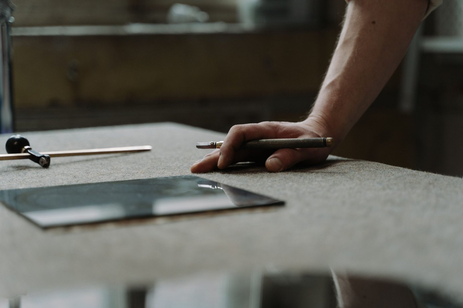 A craftsperson's hand using a glass cutter tool to score a sheet of glass in a workshop.