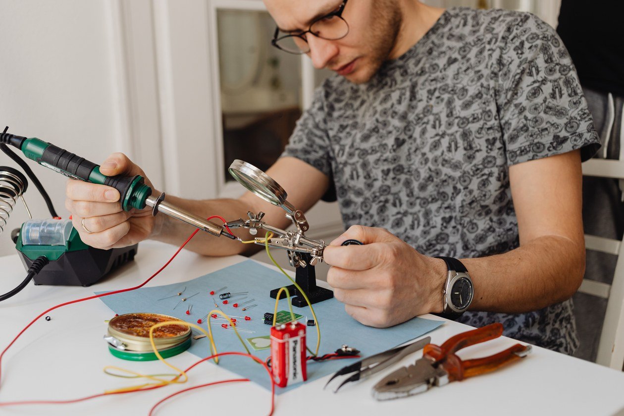 Hands holding a soldering iron with a fine wisp of smoke rising from the tip, the same grip technique used when soldering stained glass seams.